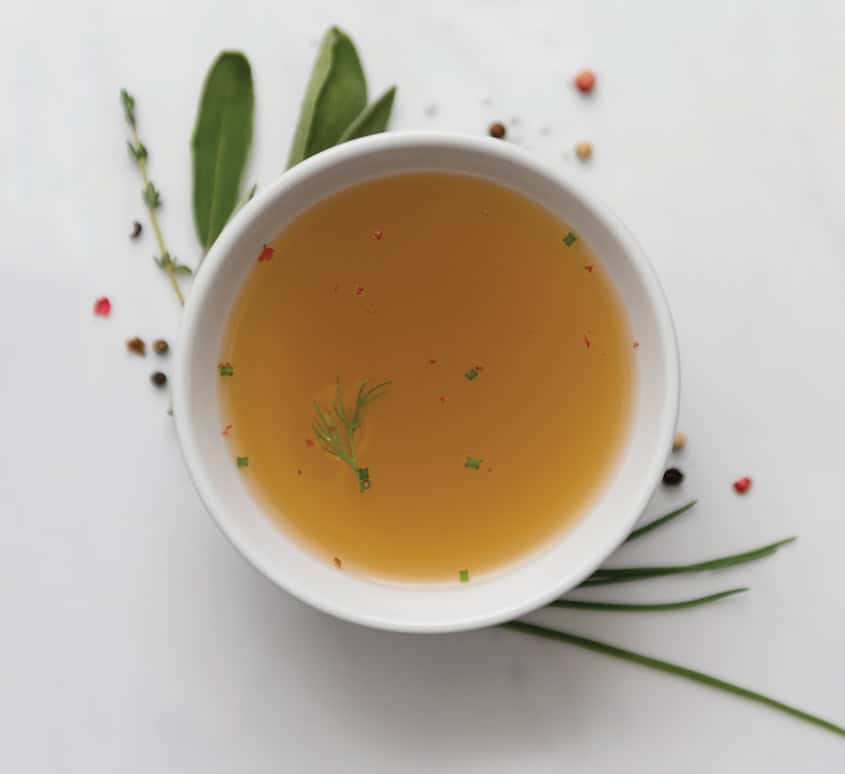 A white bowl filled with golden bone broth, garnished with fresh herbs and surrounded by scattered peppercorns and greenery, embodying the essence of nourishing collagen broth and instant bone broth powder.
